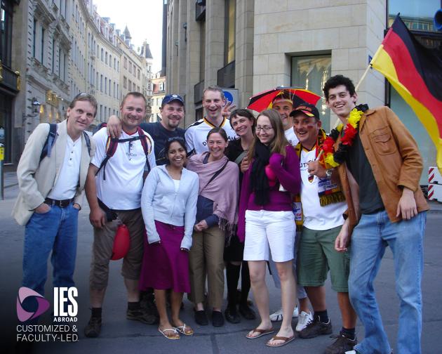 a group of students with their instructor posting with a German flag with the IES Customized & Faculty-Led logo over the photo