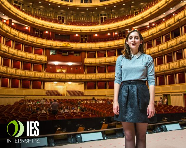 a student intern standing on stage in front of an empty auditorium with the IES Internships logo over the photo