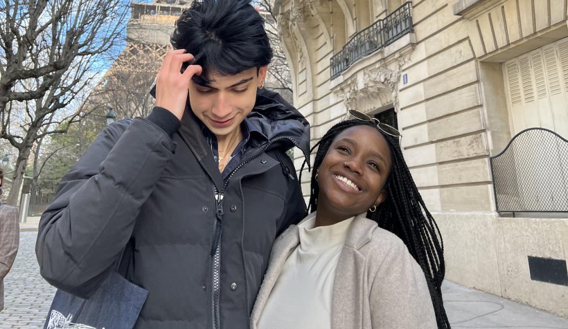 Two students, one taller than the other, are in front of the Eiffel Tower in Paris, France. One student smiles and the other is fixing his hair.