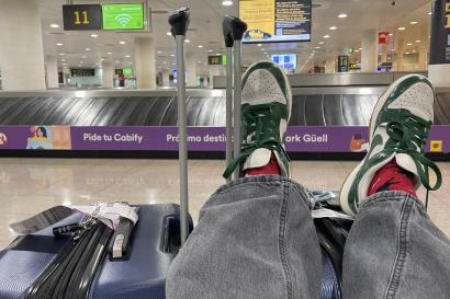 legs with black jeans and green and white Dunks rest atop of two blue suitcases in front of the baggage claim terminal at BCN airport.