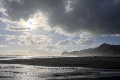 This photo shows Piha Beach during a sunset. Dark clouds are in the upper half of the photo, while the sun peaks through the empty space. The foreground shows the water illuminated by the sun and the background shows a few people along the shore.