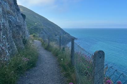 A narrow coastal path winds along a cliff edge, bordered by a fence and wildflowers. To the right, it overlooks a calm blue sea under a clear sky, with a hilly landscape on the left.