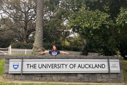 A girl wearing a University of Auckland t-shirt poses in front of the large stone sign that says The University of Auckland.