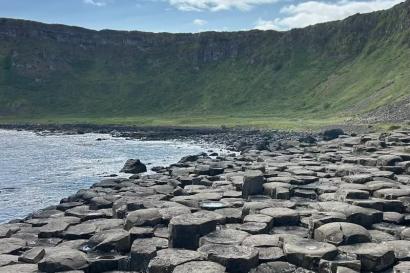 One of the many beautiful views at Giant's Causeway, including the circular rocks, coast, green hills, and blue skies with scattered clouds overheard.
