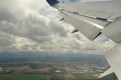 Cloudy sky view from the airplane taking off from Dublin. The wing of the airplane, cloudy blue skies, and green fields are visible.