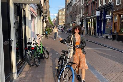 On a brick sidewalk in the streets of Amsterdam, Brooke, a white woman with brown curly hair, is pictured in an orange dress and black jacket, walking with her city bike. She is smiling at the camera.