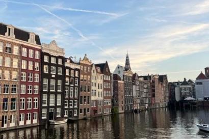 A row of tall, eloquent buildings are shown, reflected in the canal in the foreground. The sky is blue with wispy clouds high overhead. 