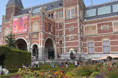 The eloquent, red facade of the Rijksmuseum is pictured, foregrounded are the gardens of the Rijks with colorful flowers and shrubbery.