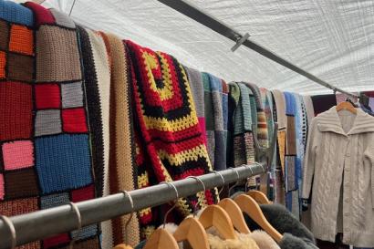 At one of the IJ-Hallen flea market tents, crocheted, colorful blankets are displayed hanging over a rack. In front of them wooden hangers bear a variety of sweaters.