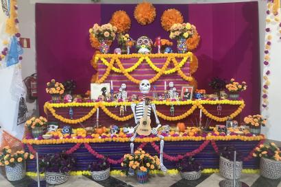 A vibrant Día de los Muertos altar from the Museo de América in Madrid. The ofrenda features marigold garlands, papel picado, skeleton figurines, candles, sugar skulls, traditional Mexican textiles, and offerings like fruits and food, all arranged on a tiered altar against a magenta backdrop.