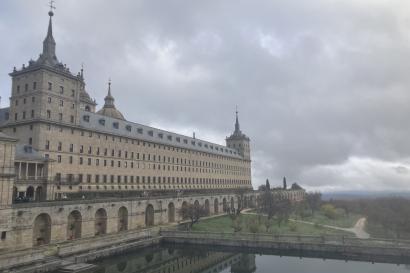 The grand exterior of El Escorial, with its tall spires and stone facade, reflected in a calm pond, surrounded by fog and autumnal trees.