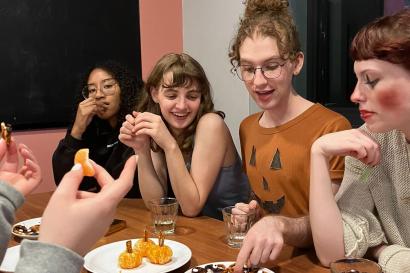 Candid photo of a group of students sitting around a long table. Some of the students are wearing Halloween costumes, and there are Halloween-themed snacks on the table. 