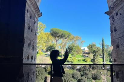 A silhouette of Brooke, a young woman with curly brown hair, is in the center of this photo, taking a photo of the Colosseum. Behind her is the bright blue sky, in an arch around Brooke.