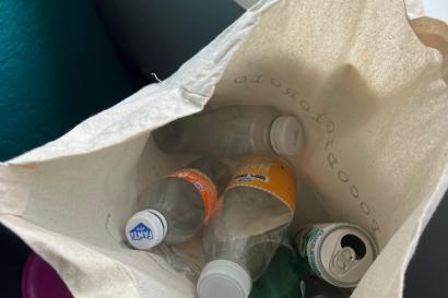 We are looking down into an open tote bag, halfway filled with empty plastic bottles and an aluminum can. 