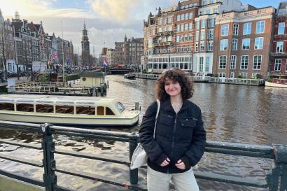 Brooke, a white woman with curly brown hair, smiles at the camera. Behind her is the Amstel river in Amsterdam, surrounded by canal houses where the Amstel meets Spui and Rokin.