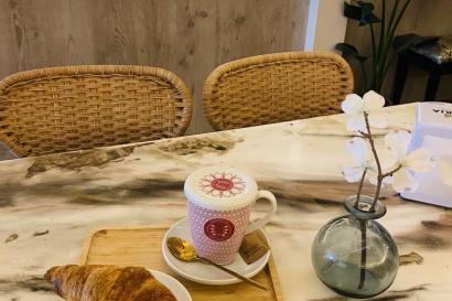 A croissant and a hot macha latte in a pink cup next to a small flower vase on a marble table