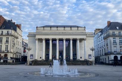 A fountain in the middle of a square, with a large neo-classic building with pillars at the far end. 