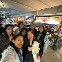 In this photo, there are seven people crowded together for a selfie at a busy night market. The girl in the front wears a light colored jacket, while another girl holds a colorful llama. The rest wear dark colored clothes. In the background, there is a booth that says "Tanghulu".