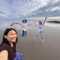 A wearing a brown shirt takes a selfie as two other girls jump in the background. The two girls jumping are wearing white shirts and jeans. The three girls are at a beach with heavy cloud cover and the sun peeking through the clouds in the background.