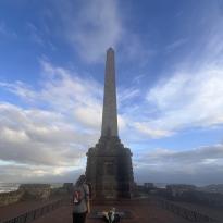 A girl stands in front of a tall, narrow monument. The sky is cloudy.