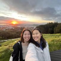 Two girls take a selfie atop Mount Eden during sunrise. The background shows a dark sky with a bright orange dot.