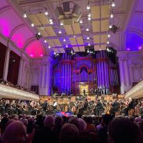A large symphony orchestra with an organ in the background. The room has pink and purple lighting.