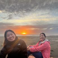 Two girls sit on the sand while the sun sets behind them. The girl on the left wears a black puffer coat and the girl on the right wears a pink jacket. The sky is orange.
