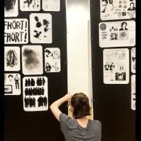 Young woman sitting on floor cross-legged, facing away from camera, securing original drawings to gallery wall 