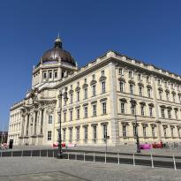 An imposing building is in the forefront, with a clear blue sky behind it. 