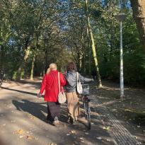 In Vondelpark, a large park in Amsterdam, the backs of Brooke, a white woman with curly brown hair, and her friend Vicki, a while woman with blonde hair and a red coat, are pictured---they walk together, Brooke walking her bike alongside them.