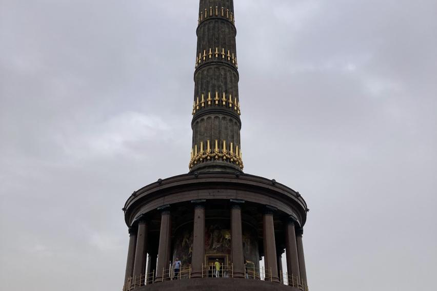 A view looking up at the Berlin Victory Column against a cloudy, gray sky. The golden goddess of victory shines at the very top, and gilded cannons line the sandstone column in rows. The tower sits atop a round base made of polished red granite, which itself is held up by a ring of columns.