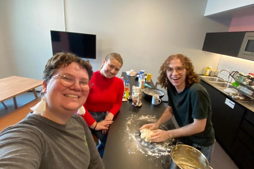 Selfie of three students around a cluttered counter in a communal kitchen. A space has been cleared on the counter, where one of the students is kneading dough.