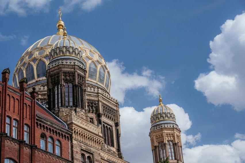 Photograph of the elaborately decorated dome of the Neues Synagogue in Berlin, with a cloudy, blue sky in the background. 