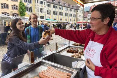 two students smiling at a wurst stand with a vendor behind glass handing them a bun with a sausage