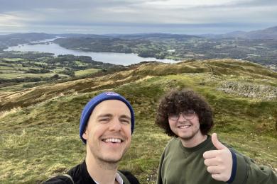 two students standing holding a thumbs up in front of rolling grass hills