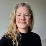 Woman with long blonde hair and glasses in front of a grey wall 