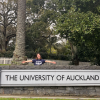 A girl wearing a University of Auckland t-shirt poses in front of the large stone sign that says The University of Auckland.