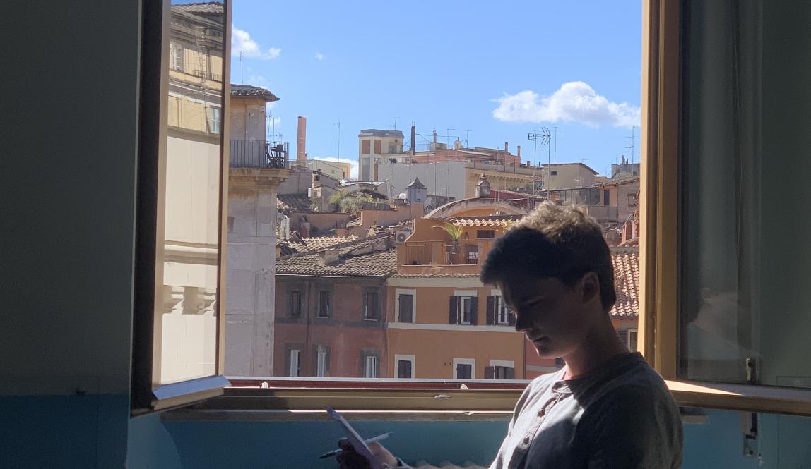 A student at the Rome Center works on their assignment with the window open. Outside the window are terracotta-colored buildings.