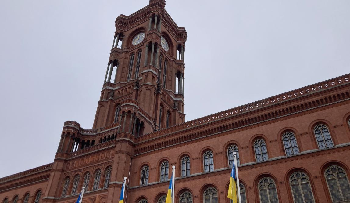 Image of the Rotes Rathaus in the Berliner Innenstadt. The Neo-Renaissance redbrick town hall is set against a grey, cloudy sky. A row of Ukrainian flags on flagpoles are lined up in front of the building.