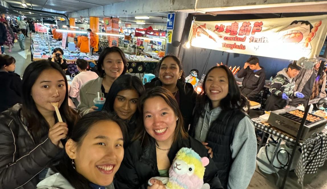 In this photo, there are seven people crowded together for a selfie at a busy night market. The girl in the front wears a light colored jacket, while another girl holds a colorful llama. The rest wear dark colored clothes. In the background, there is a booth that says "Tanghulu".