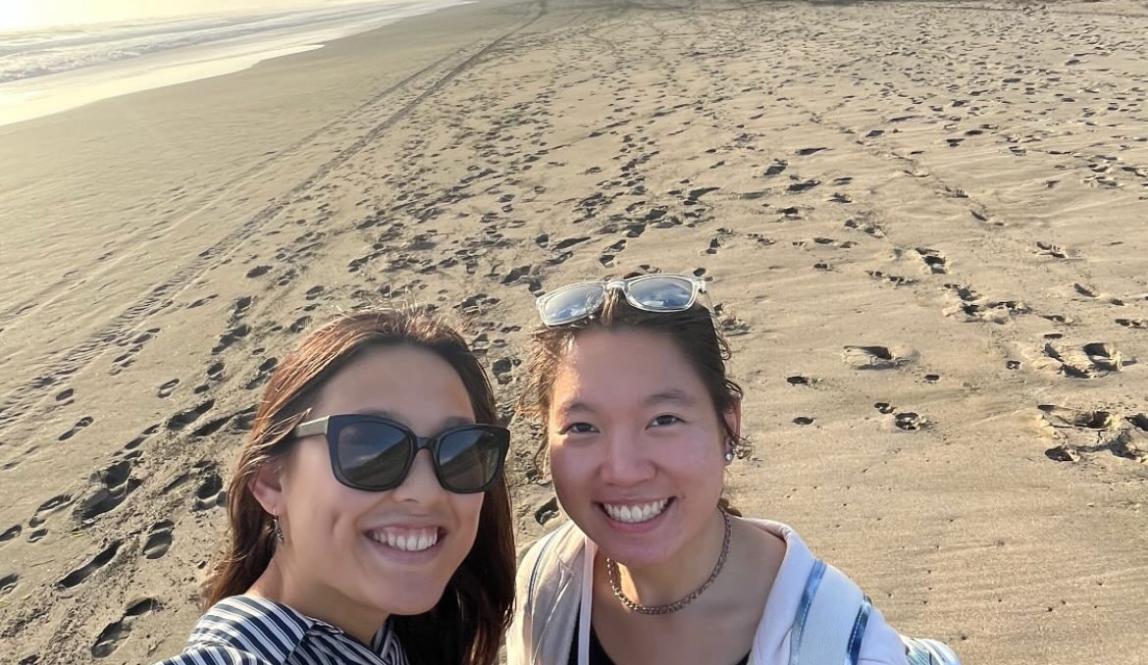 In this photo, there are two girls posing for a selfie at the beach. The girl on the left wears a striped shirt with sunglasses, while the girl on the right wears a black tank top and black pants. There are thick clouds overhead and the sun is setting in the background.