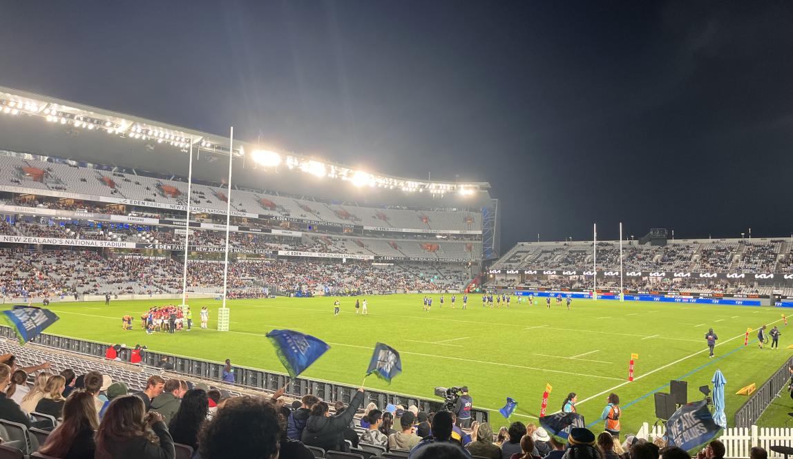 This photo shows a rugby match. In the foreground, there is a crowd that waves blue flags, and in the background there are several rugby players on the green field. There are also bright lights in the background. 