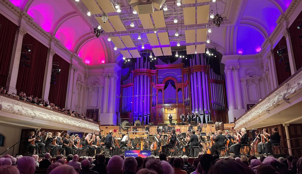 A large symphony orchestra with an organ in the background. The room has pink and purple lighting.