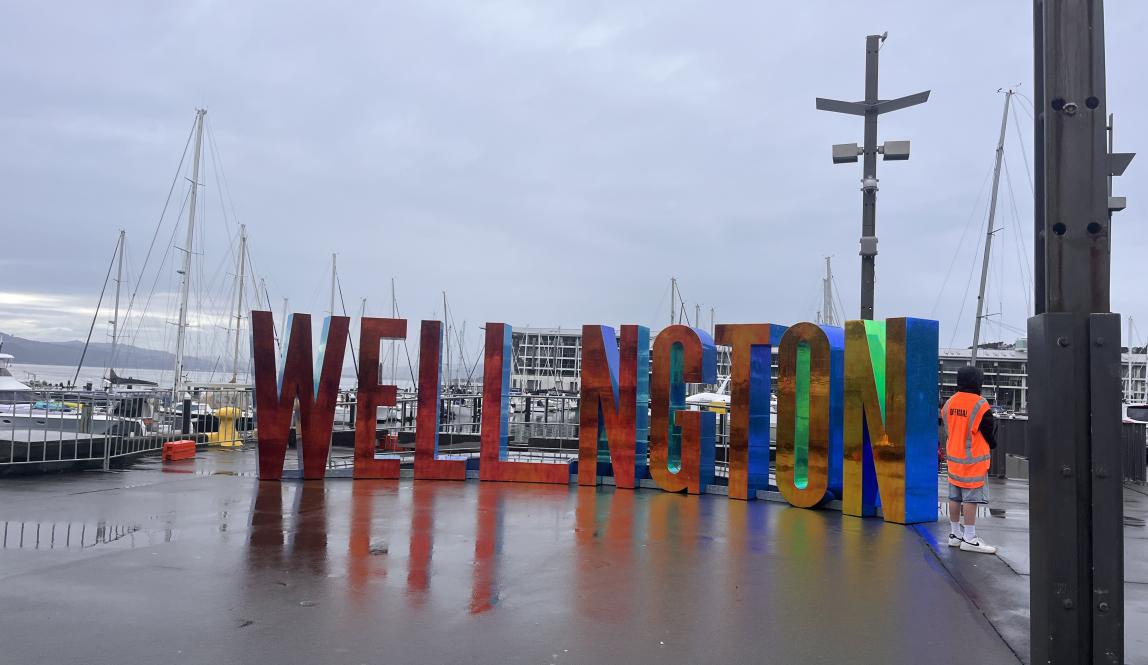 Large block letters spell out the word Wellington at a harbor. The letters are solid and sculptural-like.
