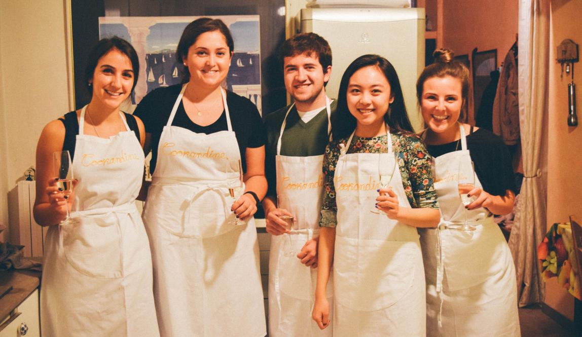 Five students in aprons at a cooking class, holding glasses of wine and smiling.