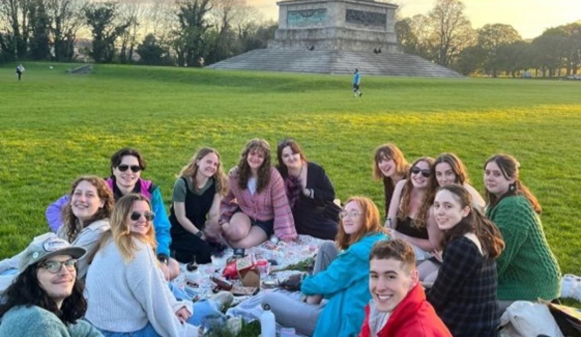 group of students sitting on the grass with picnic stuff in front of a stone monument