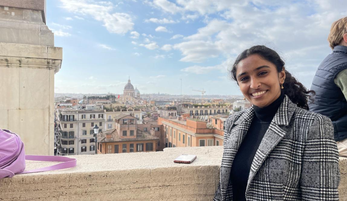 A student in a grey houndstooth blazer smiles from a terrace with the city of Rome sprawling behind her.