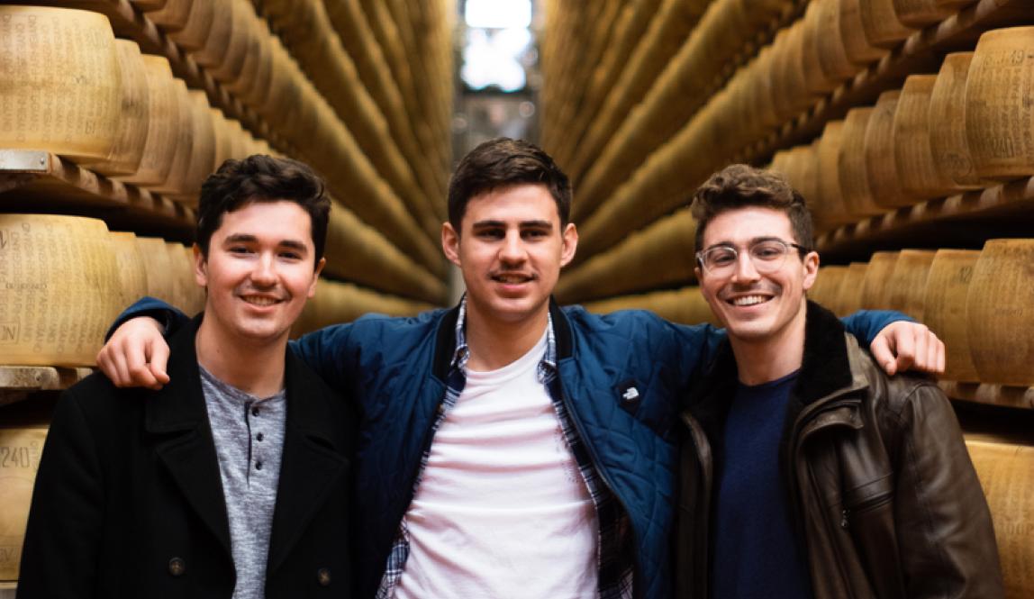 Three friends stand in front of a wall of aging cheese wheels, smiling.