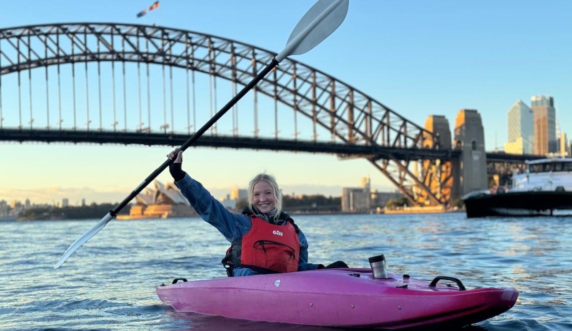 a student posing from a kayak in Sydney Harbour, holding their paddle in the air above their head in celebration