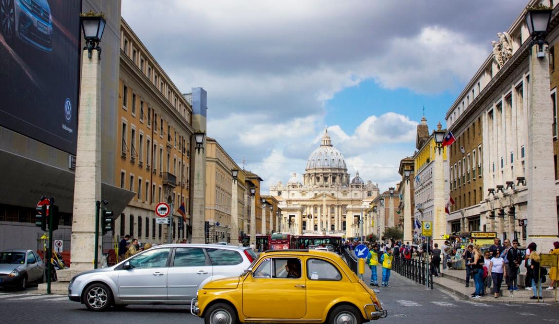 A vintage yellow car passes by on a busy street in front of the Vatican.